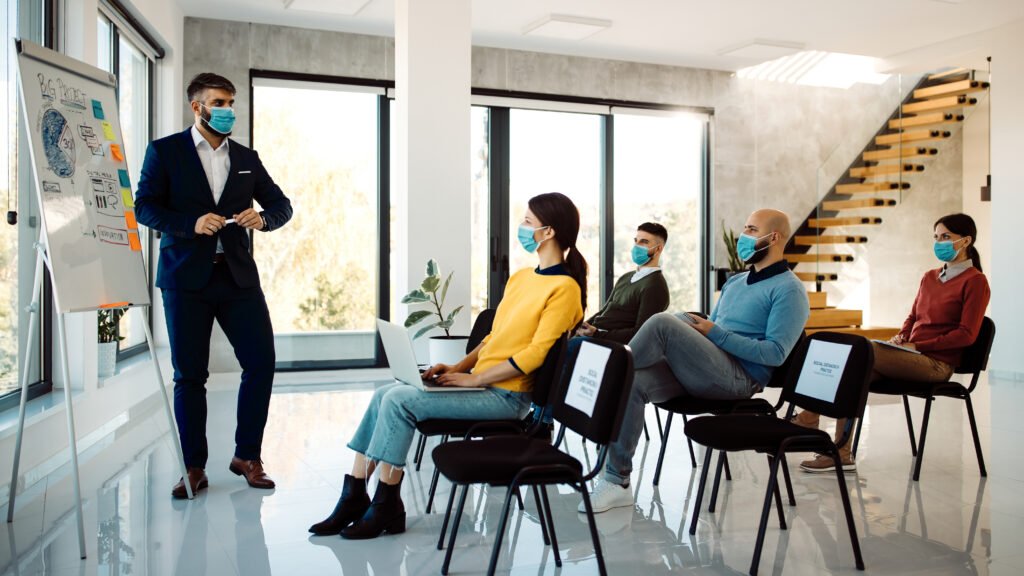 Male entrepreneur talking to group of people during a business seminar in board room. All of them are wearing face masks due to COVID-19 pandemic.